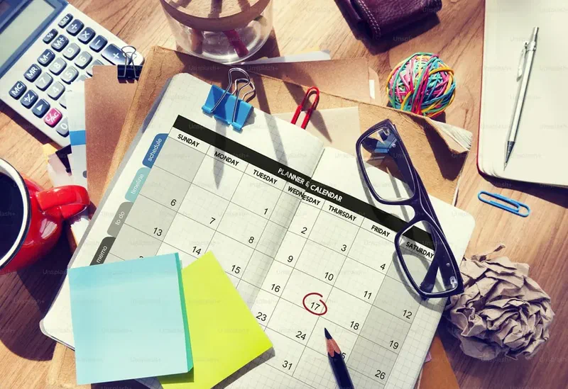 Monthly planner calendar with a marked deadline on a cluttered office desk with stationery and glasses