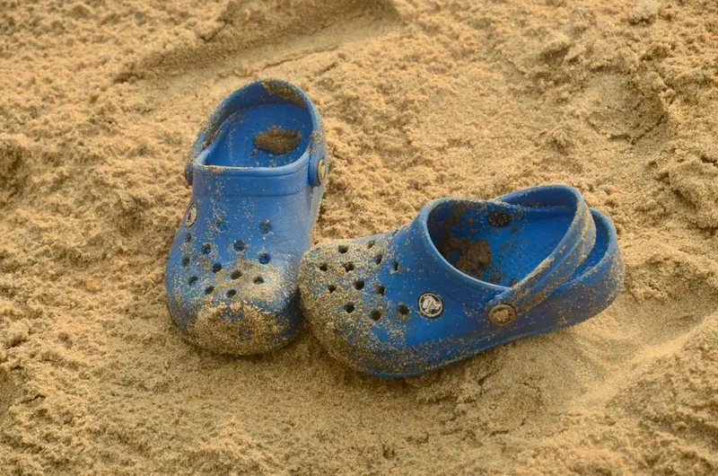 Pair of blue clogs partially covered with sand on a sandy beach.