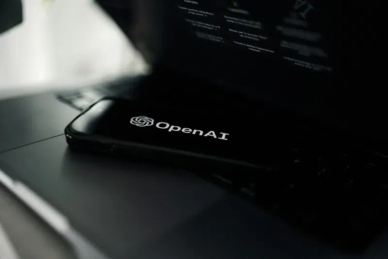 Smartphone displaying the OpenAI logo placed on a laptop keyboard with an AI interface visible in the background.