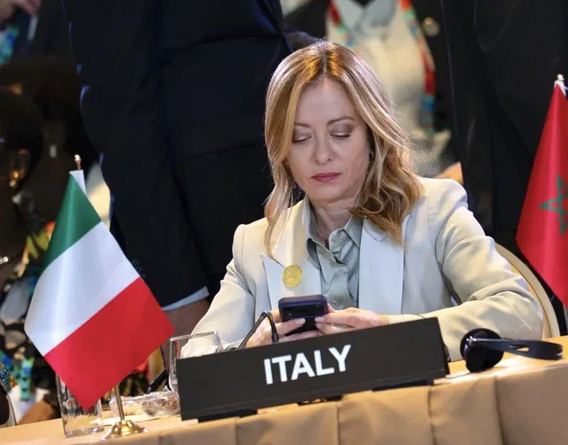 Italian Prime Minister seated at an international summit, using a smartphone beside the Italian flag and an Italy nameplate.